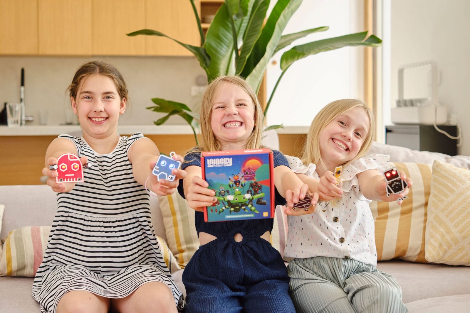 Three children sit smiling on the sofa and hold the Dusty the Wacky Robot educational set.